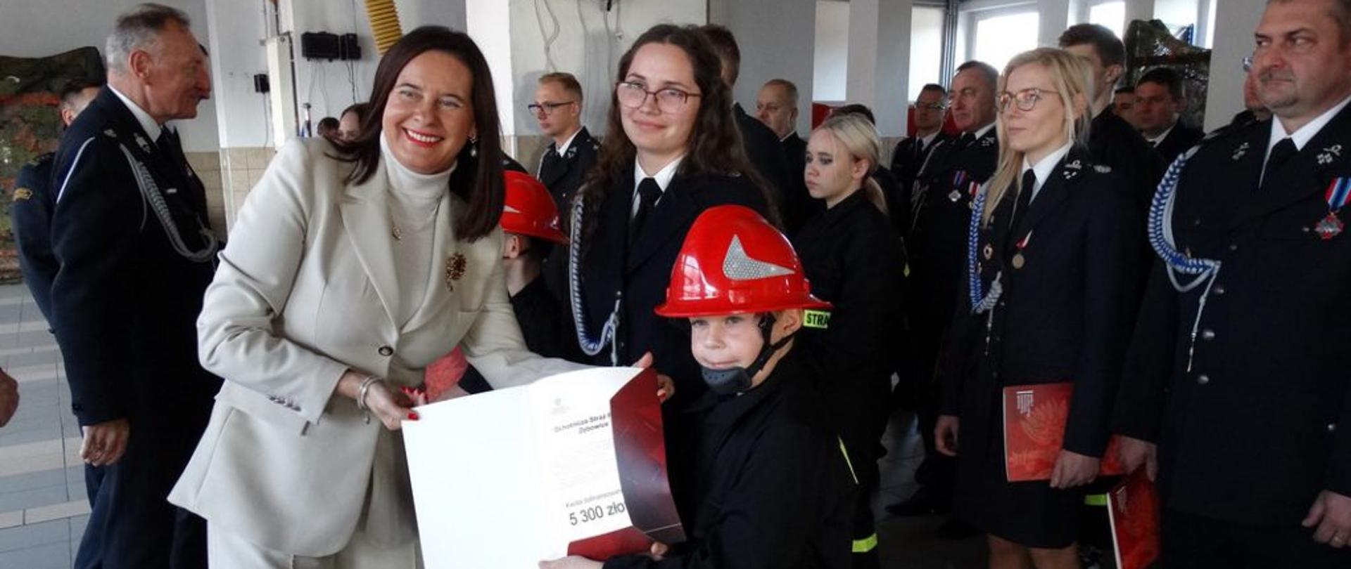 Strażacy z OSP w mundurach wyjściowych i MDP w czerwonych hełmach otrzymują promesy