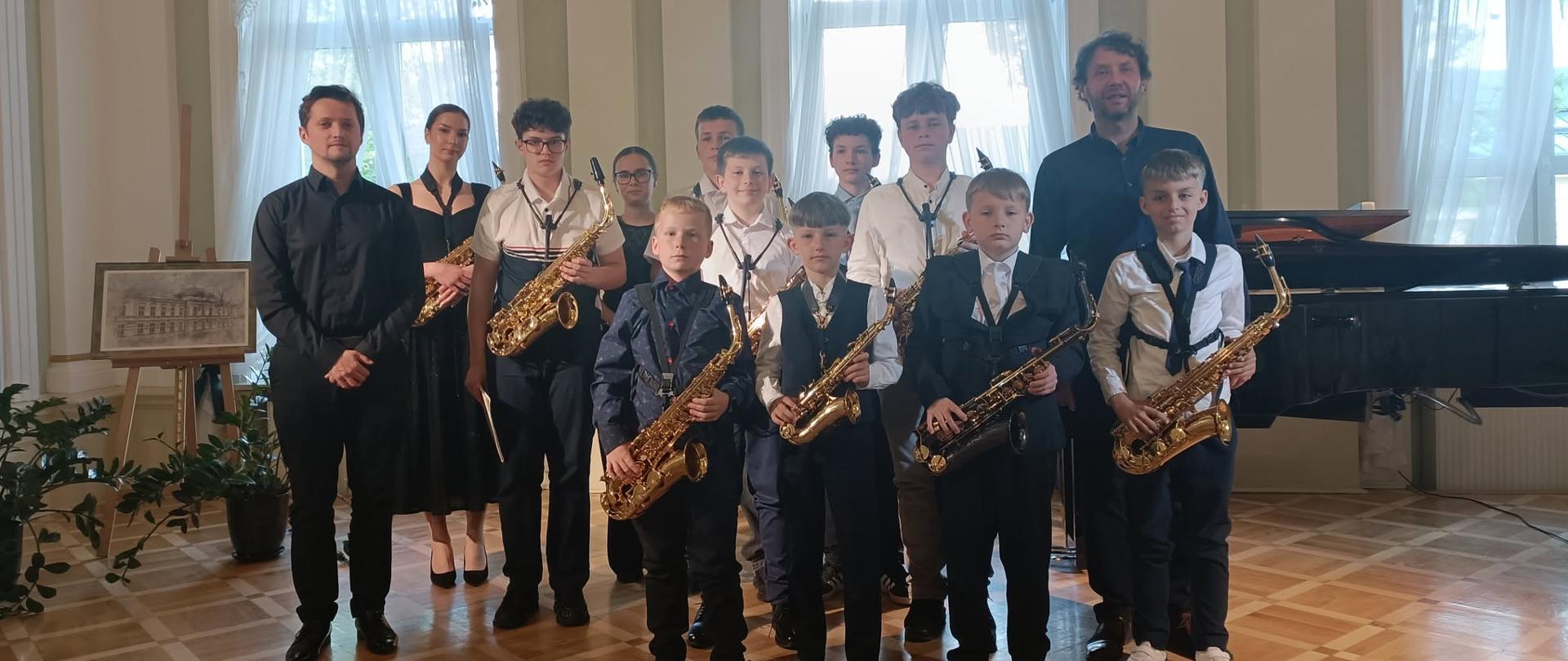 djęcie przedstawia występ muzyczny młodego akordeonisty w jasnym pomieszczeniu na poddaszu ze skosami i oknami dachowymi.
Pośrodku pokoju, na jasnej podłodze z paneli, siedzi bokiem do obiektywu chłopak ubrany w elegancką czarną koszulę, ciemne spodnie oraz czarne buty. Gra on na dużym, czarnym akordeonie guzikowym, z dużym skupieniem patrząc w stronę instrumentu. Przed muzykiem, po prawej stronie kadru, ustawiony jest czarny, metalowy pulpit z rozłożonymi nutami.
W tle bezpośrednio za akordeonistą widoczna jest biała, harmonijkowa ścianka dekoracyjna ozdobiona pofalowanymi, czarnymi liniami. Po lewej stronie, w głębi sali, stoi duży, lśniący czarny fortepian skrzydłowy z podniesioną klapą. Ściana w tle nad fortepianem została udekorowana papierowymi, okrągłymi rozetami w kolorach zielonym, różowym i czerwonym, co podkreśla popisowy i odświętny charakter wydarzenia.
