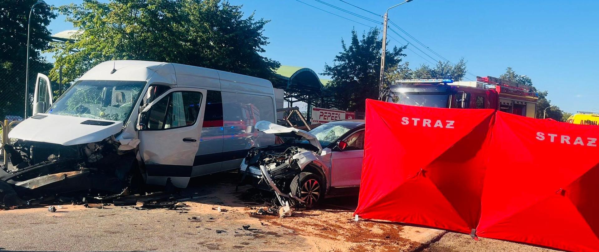 dwa rozbite auta – samochód typu bus oraz samochód osobowy znajdujące się na drodze. Z prawej strony rozłożony strażacki parawan. W tle widoczny samochód pożarniczy. 