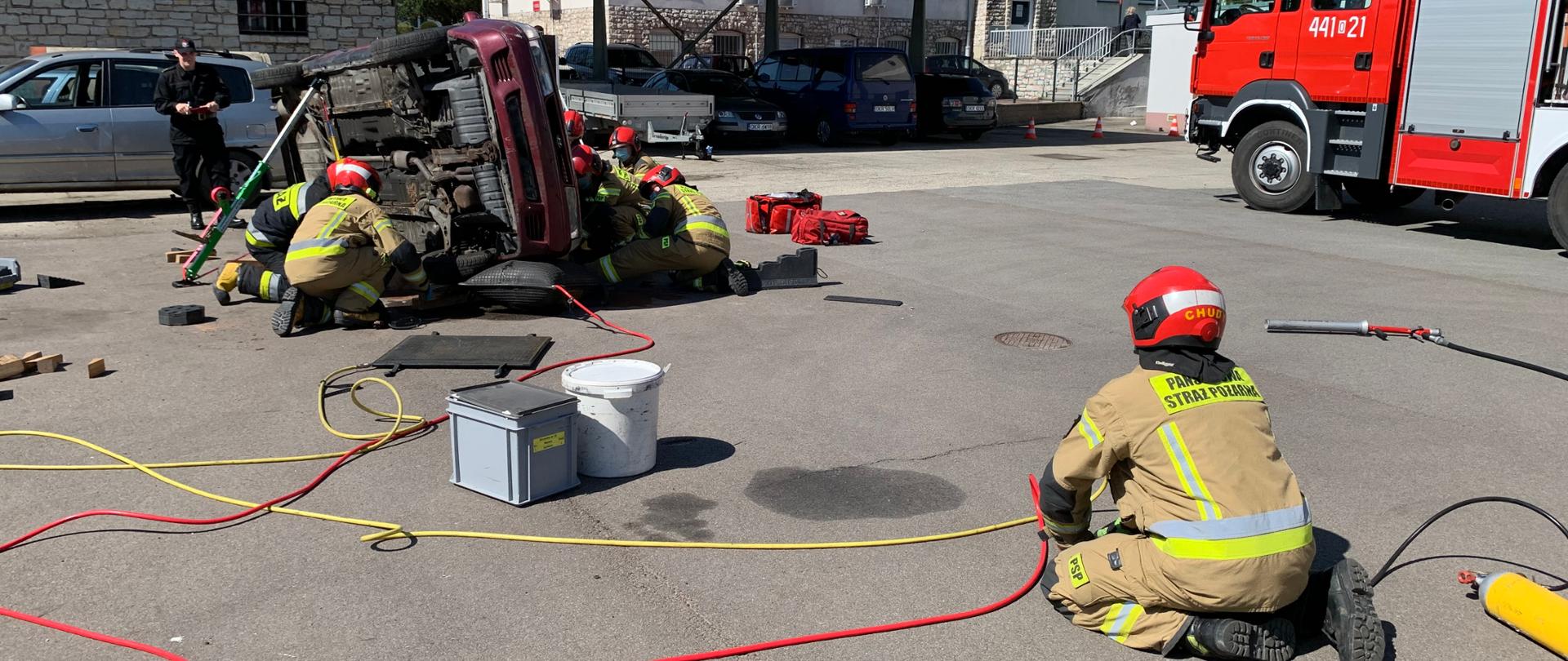 Zdjęcie przedstawiające symulowaną akcję ratowniczą. Na pierwszym planie tyłem do patrzącego klęczy na ziemi strażak obsługujący zestaw pneumatyczny. Strażak spogląda na przewrócony na lewy bok samochód osobowy, który podniesiony został za pomocą poduszki pneumatycznej. Jedna z poduszek leży przed samochodem gotowa do użycia. Powietrze do poduszek doprowadzone jest przewodami w kolorach żółtym i czerwonym. Samochód osobowy z lewej strony podparty jest podporą ratowniczą. Przy pojeździe klęczą strażacy (5) którzy zabezpieczają proces podnoszenia i wydobycie poszkodowanego spod auta. Za pojazdem stoi oficer oceniający pracę ratowników w obraniu koszarowym. Ratownicy w ubraniach specjalnych koloru piaskowego, jeden w granatowym. Wszyscy w czerwonych hełmach . W tle pobliskie zabudowania i wiata. Po prawej uwidoczniono przednią część samochodu pożarniczego – lewy bok samochodu. 