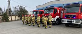 Strażacy stojąc na baczność na tle samochodów gaśniczych oddają cześć poległym strażakom z Ukrainy.
