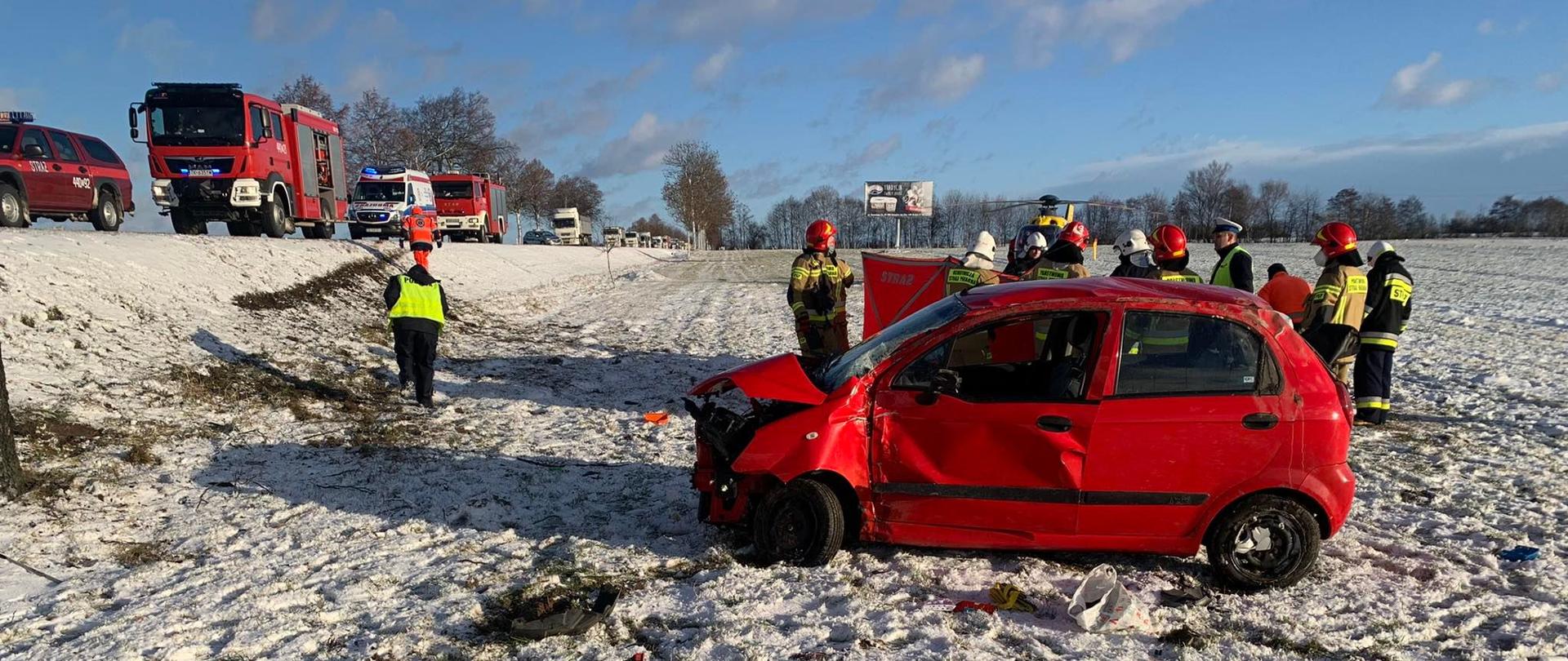 Zdjęcie przedstawia samochód osobowy koloru czerwonego oraz parawan koloru czerwonego przy, którym znajdują się strażacy, ratownicy LPR oraz funkcjonariusz Policji. W tle widoczny śmigłowiec LPR koloru żółtego, samochody straży pożarnej koloru czerwonego oraz karetkę Państwowego Ratownictwa medycznego koloru białego. W oddali widoczny radiowóz Policji koloru szarego, oraz samochody ciężarowe na drodze.
