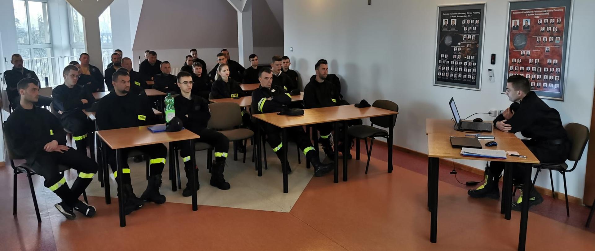 Na zdjęciu widać siedzących w sali ok. 24 osób. Wszyscy siedzą w ławkach wszyscy ubrani są w czarne strażackie stroje koszarowe. Na przeciw nich siedzi przy biurku oficer prowadzący wykłady.