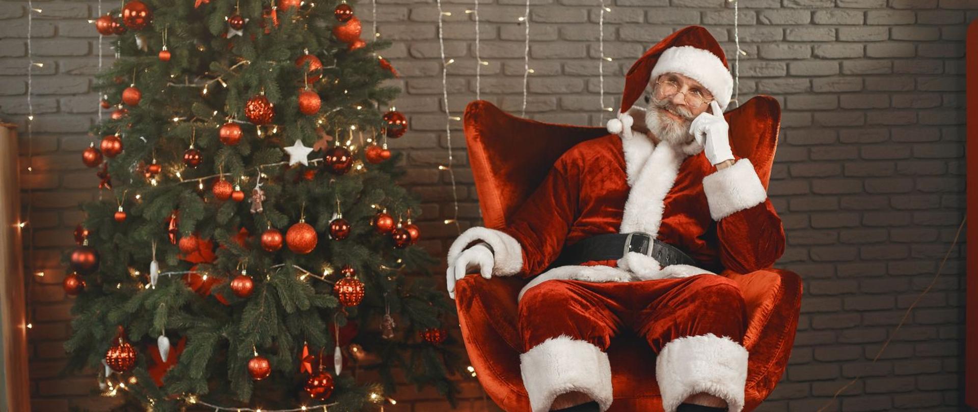 zdjęcie przedstawia po lewej przystrojoną czerwonymi bombkami choinkę, po lewej postać siedzącego w fotelu świętego mikołaja