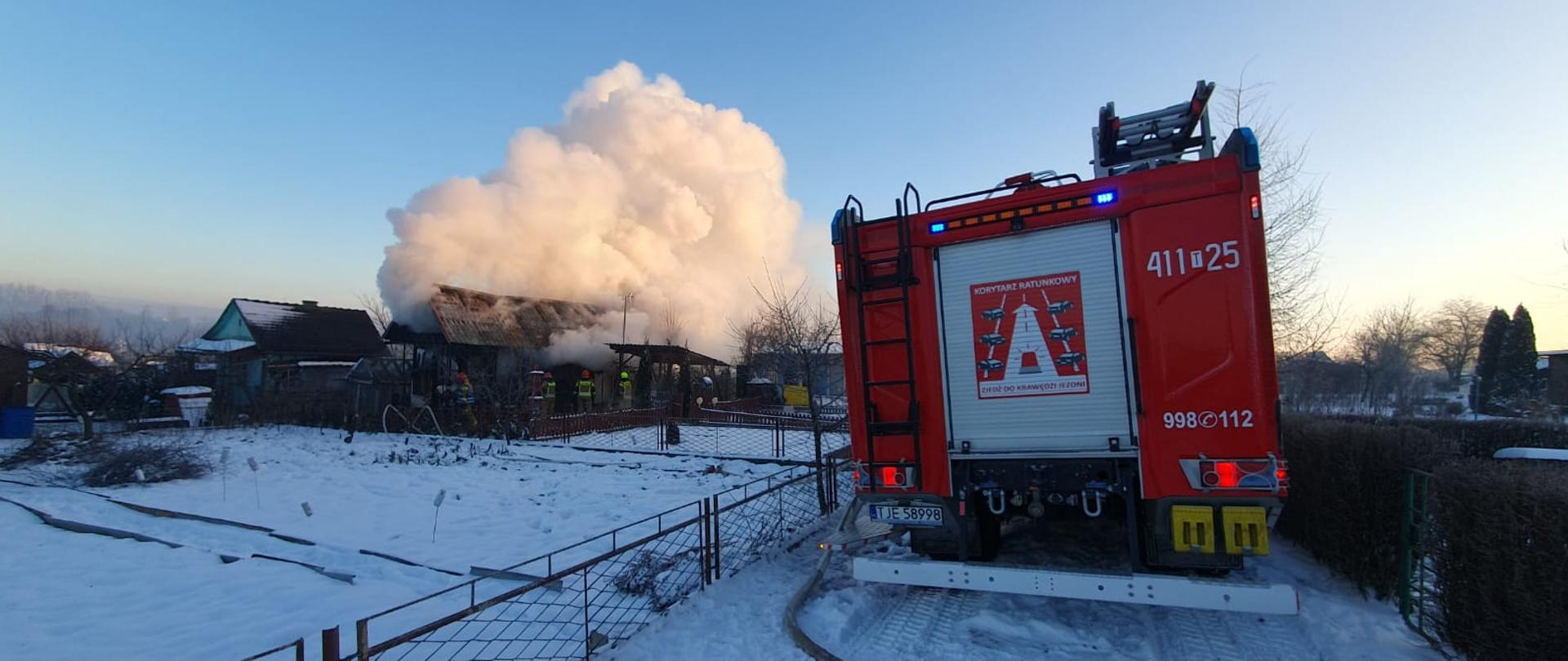 Zdjęcie przedstawia chmurę dymu unoszącą się nad drewnianą altaną letniskową na ogródkach działkowych. Na pierwszym planie tył samochodu pożarniczego.