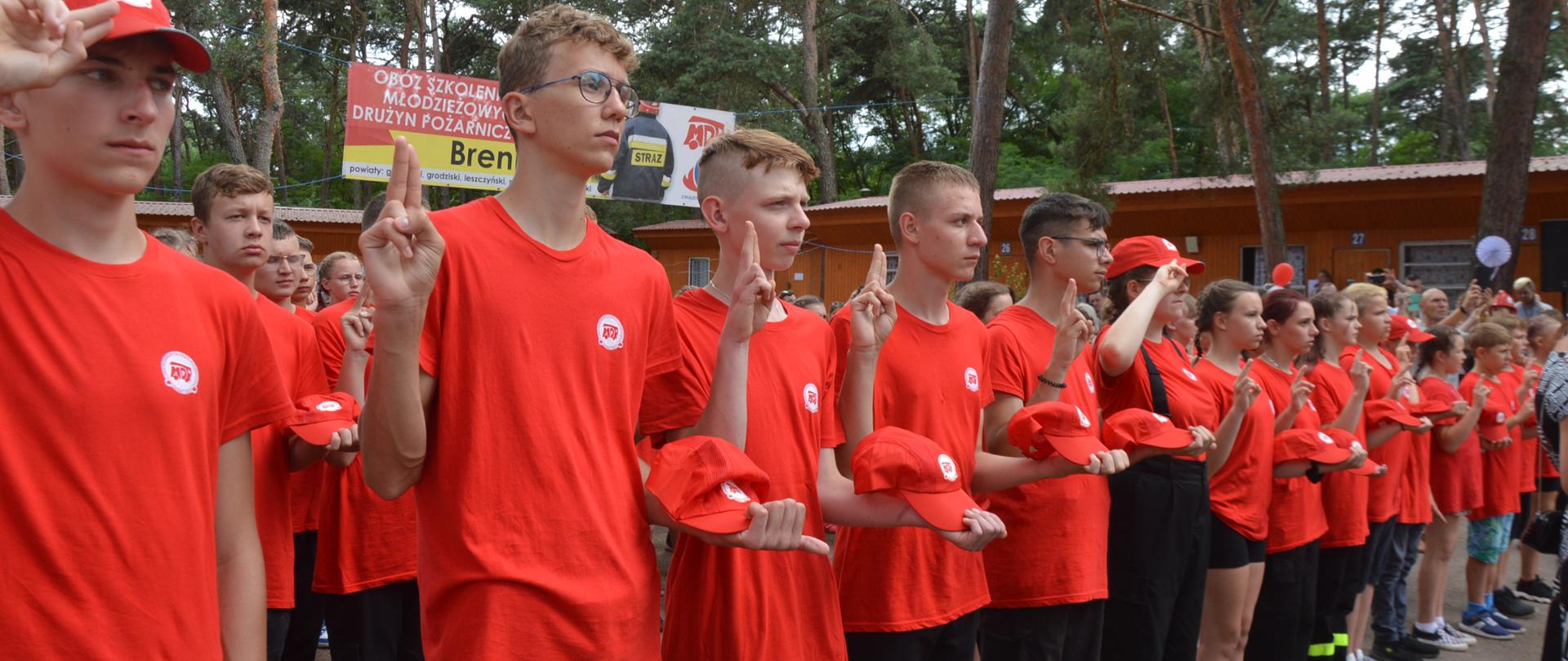 Obóz MDP Brenno. Na placu apelowym trwa uroczyste ślubowanie. Druhny i druhowie ślubujący, stojący w szeregach maja na sobie czerwone koszulki i czapki. Każdy z nich prawą rękę ma uniesioną, a w lewej dłoni trzyma czapkę. 