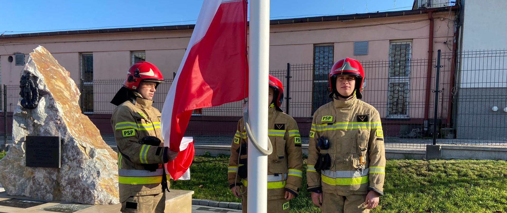 Zdjęcie przedstawia trzech strażaków: dwóch stoi na baczność, trzecie przypina flagę Polski na maszt.
