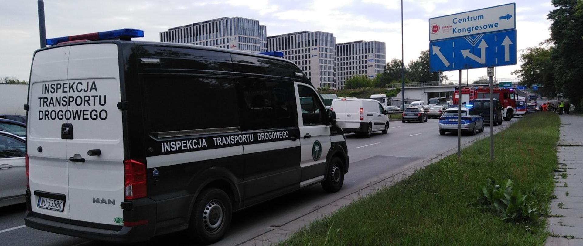 Na pierwszym planie radiowóz ITD typu furgon. W tle miejsce zdarzenia drogowego z udziałem samochodu osobowego i motorowerzysty.