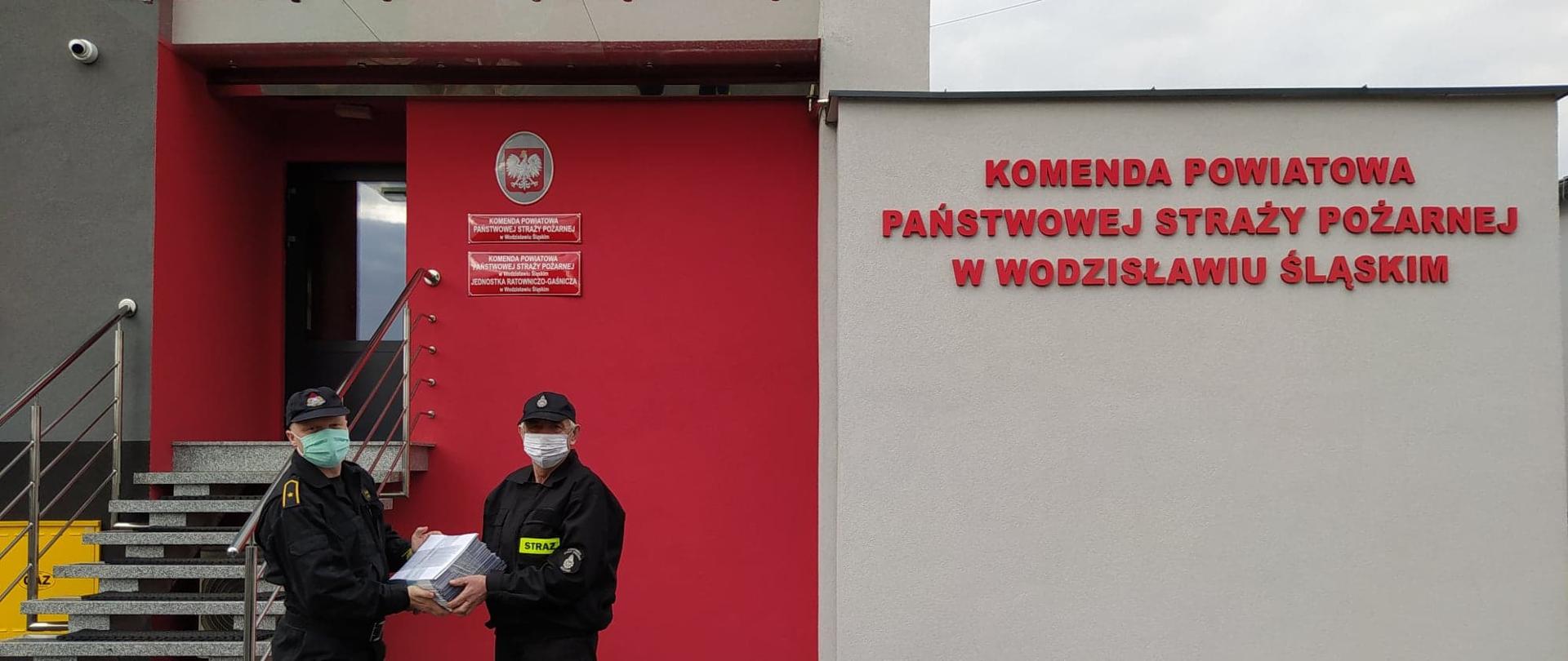 Na zdjęciu znajdują się dwaj strażacy na tle budynku komendy pozujący do zdjęcia