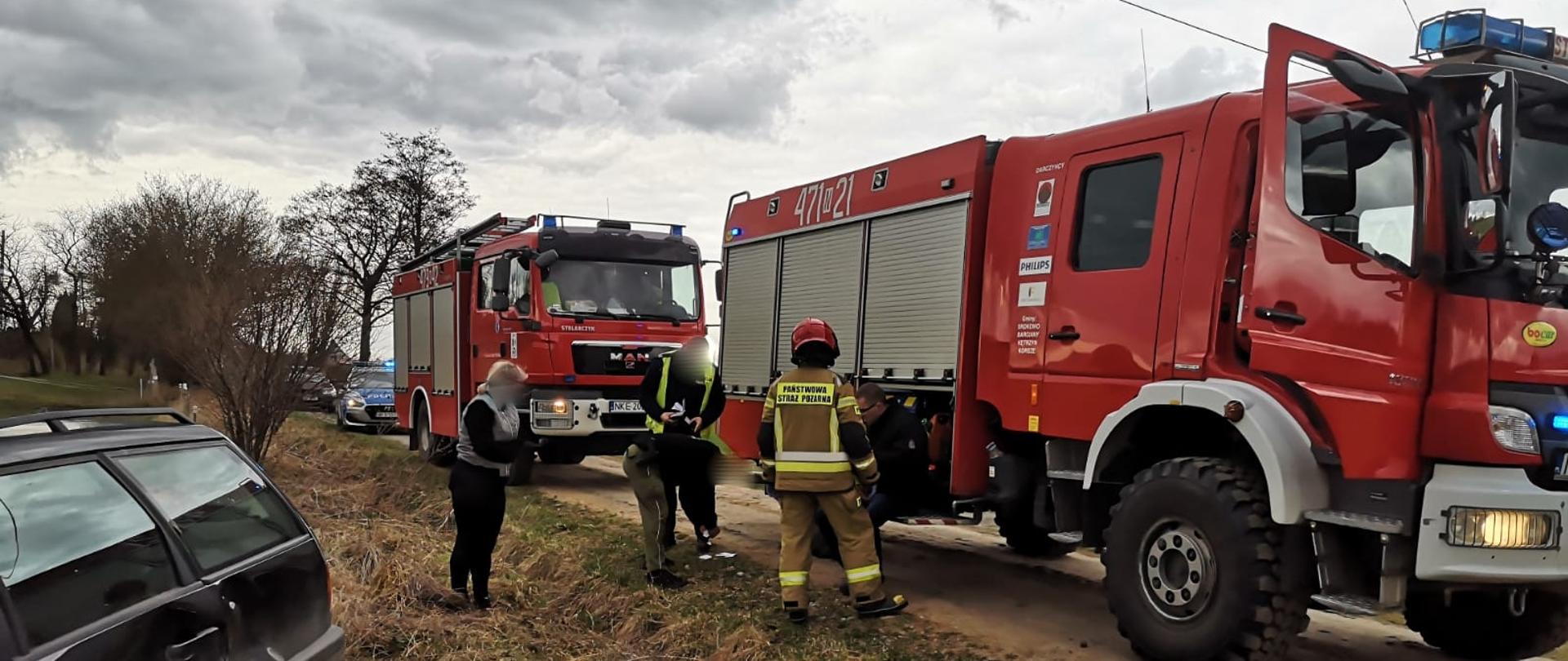 Zdjęcie przedstawia miejsce zdarzenia. Dwa czerwone wozy strażackie, samochód policji oraz uszkodzony samochód osobowy koloru czarnego. Strażak ubrany w piaskowe ubranie specjalne oraz czerwony hełm stoi przy poszkodowanym ubranym w czarną kurtkę. Obok stoi funkcjonariusz policji, straży granicznej oraz pani. 