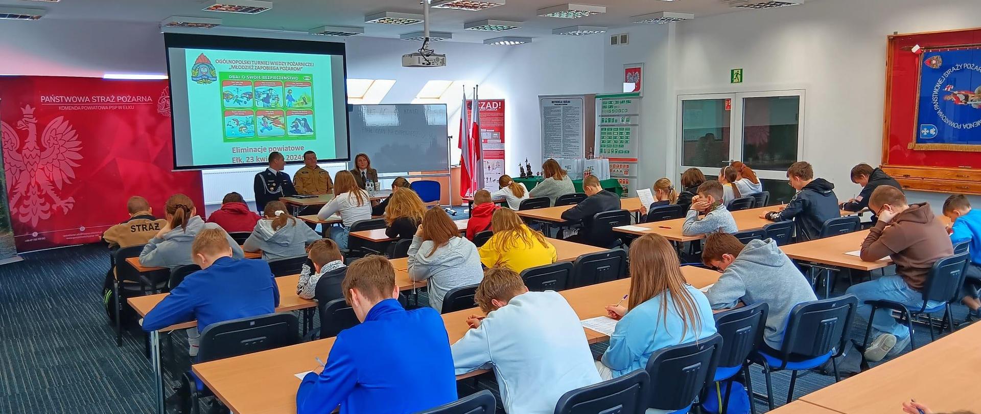 Uczestnicy w sali konferencyjnej siedzą w ławkach i piszą test