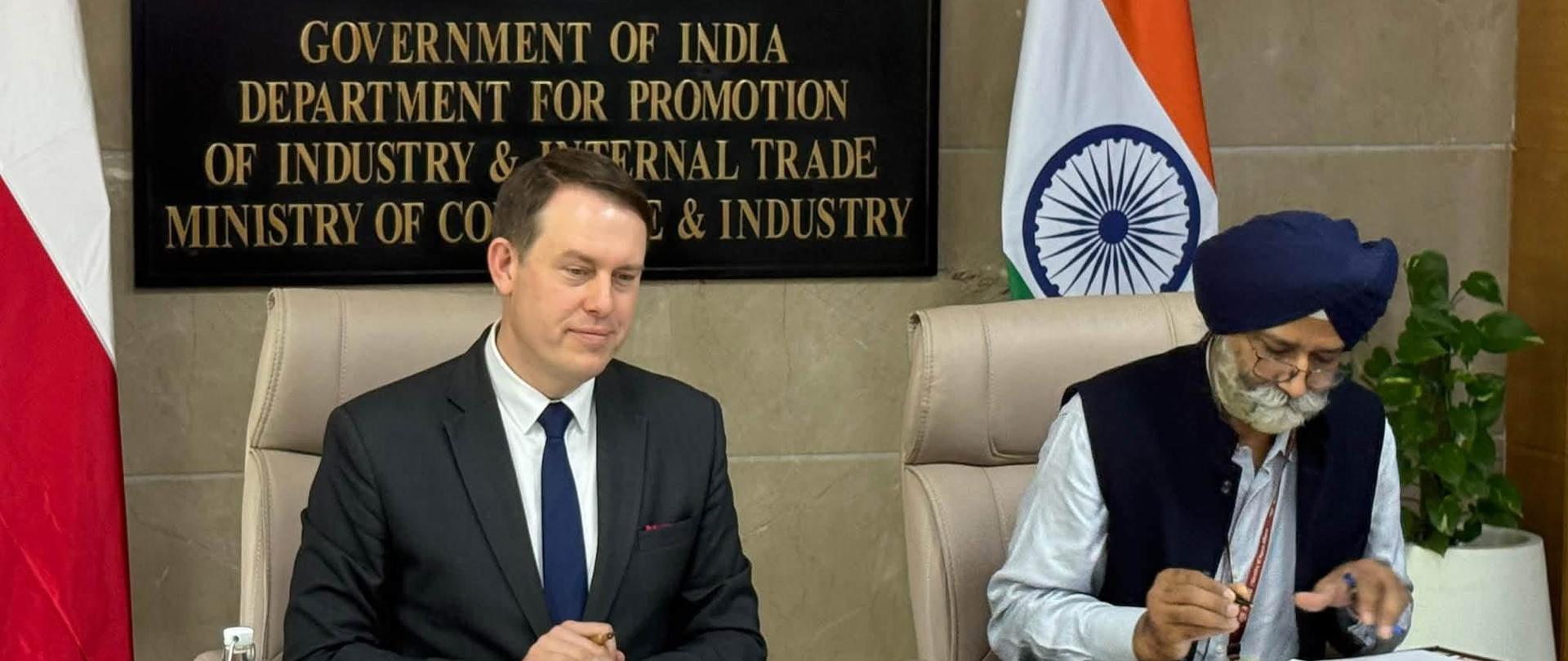 Wiceminister rozwoju i technologii Michał Baranowski pozuje z przedstawicielami strony indyjskiej po 7. posiedzeniu Polsko-Indyjskiej Komisji Współpracy Gospodarczej w Nowym Delhi. Za uczestnikami widoczne są flagi Polski i Indii.