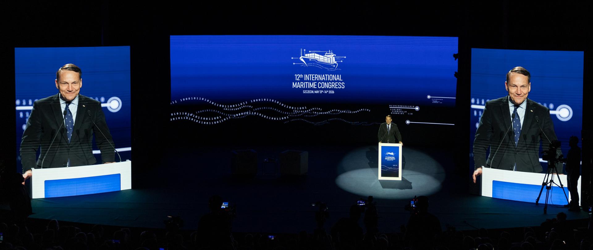 Wicepremier Sikorski stoi przy mównicy na scenie podczas 12. Międzynarodowego Kongresu Morskiego w Szczecinie. Za nim znajduje się duży ekran z grafiką wydarzenia po angielsku, a po bokach wyświetlane są zbliżenia mówcy.