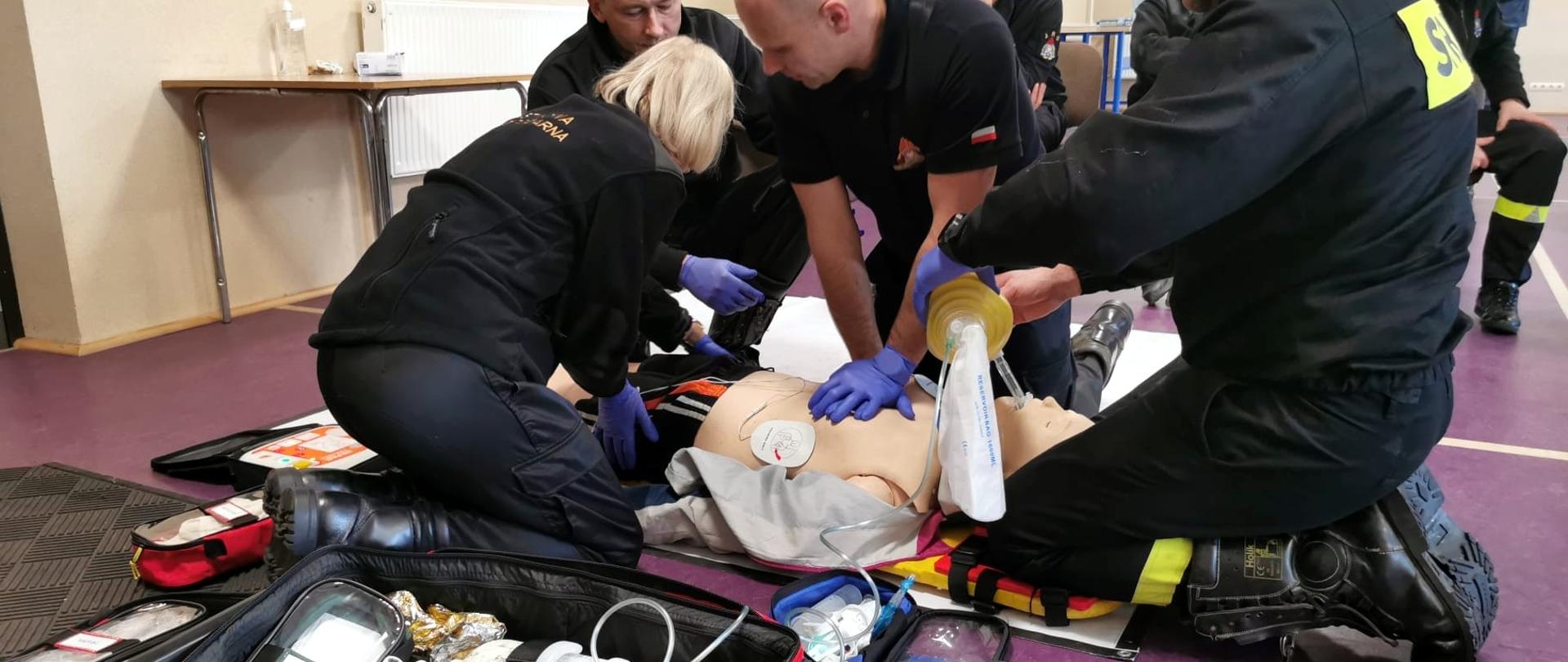 Zdjęcie zrobione podczas szkolenia z udzielania pierwszej pomocy. Na zdjęciu manekin leżący na podłodze. Strażacy ćwiczę udzielanie pierwszej pomocy. Obok leży torba medyczna.