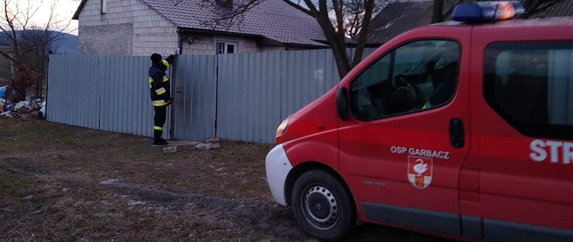 Strażacy roznoszą ulotki do gospodarstw domowych na terenie powiatu ostrowieckiego. Na zdjęciu widać strażaka przed domem mieszkalnym, który zostawia broszurę informacyjną.