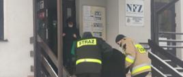 strażacy trzymają pod rękę starszą kobietę pomagają jej wejść po schodach do ośrodka zdrowia