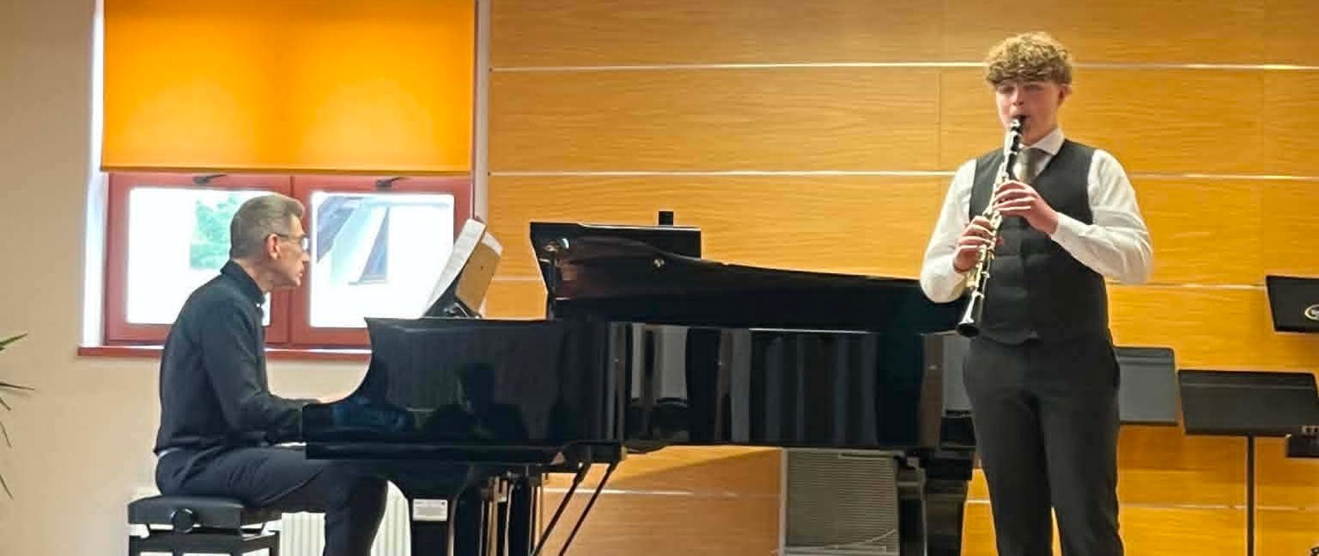 Po lewej stronie mężczyzna siedzi przy czarnym fortepianie skrzydłowym i gra na instrumencie. Jest ubrany w ciemną koszulę i ciemne spodnie. Po prawej stronie stoi młody muzyk grający na klarnecie. Ma na sobie elegancki strój galowy: białą koszulę, ciemną kamizelkę, krawat oraz ciemne spodnie. W tle znajduje się ściana wyłożona jasnym drewnem oraz okno z pomarańczową roletą. Na pierwszym planie widoczne są tyły rzędów czarnych krzeseł dla publiczności.