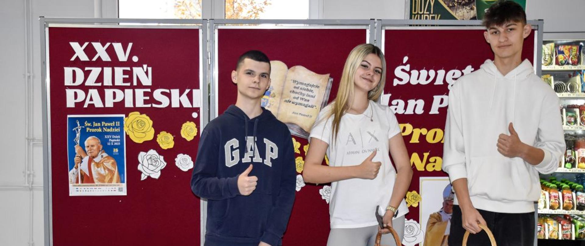 Trójka uczniów przed tablicą poświęconą Dniu Papieskiemu, w rękach mają koszyki z karteczkami (cytatami z Jana Pawła II)