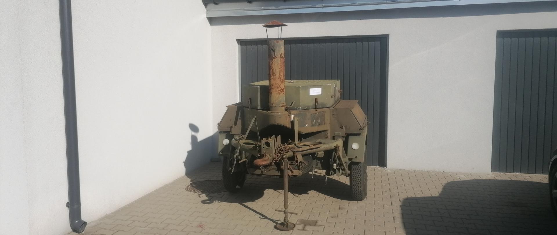 Na zdjęciu widnieje wojskowa kuchnia polowa. Kuchnia jest usytuowana na tle szarej bramy garażowej oraz kremowego budynku gospodarczego Komendy Powiatowej PSP w Zgierzu. 