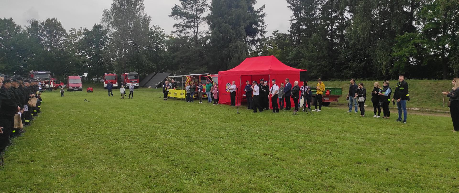 Na stadionie na tle czerwonego namiotu stoją strażacy.