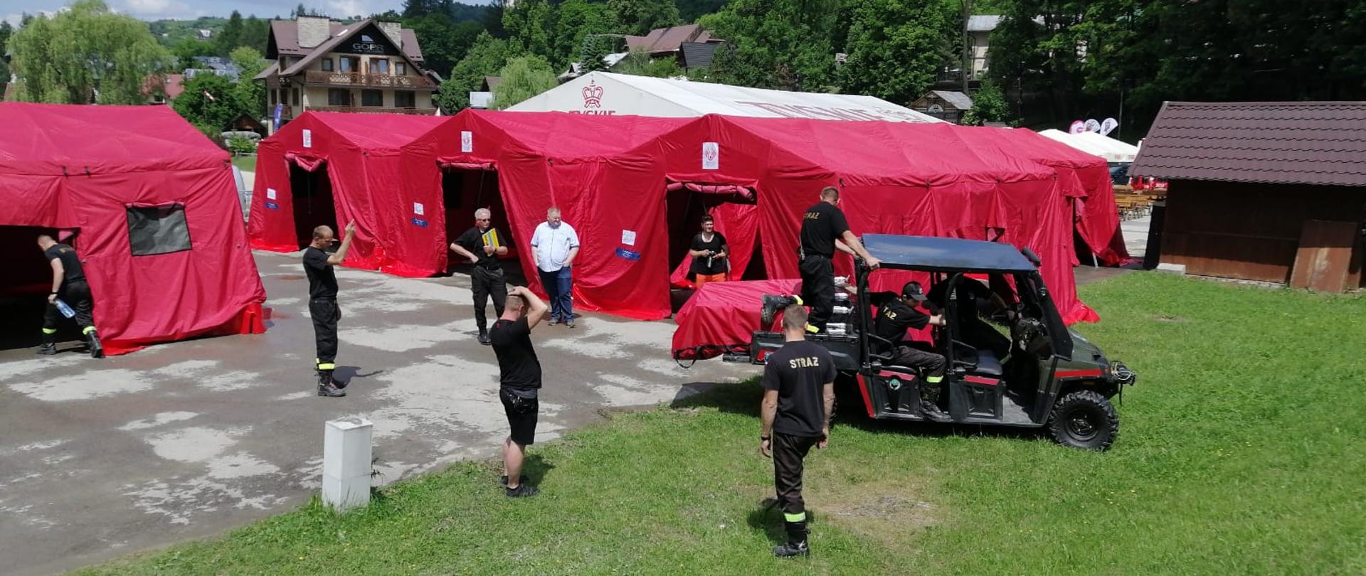 na zdjęciu cztery namioty stelażowe rozstawione na placu pod koleją linową w Szczawnicy, strażacy przy pomocy quada przewożą kolejne namioty które posłużą jako plenerowy punkt szczepień