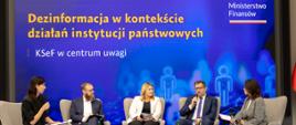 Kobiety i mężczyźni siedzą na fotelach. Rozmawiają. W tle niebieska ściana z napisem: Dezinformacja w kontekście działań instytucji państwowych.