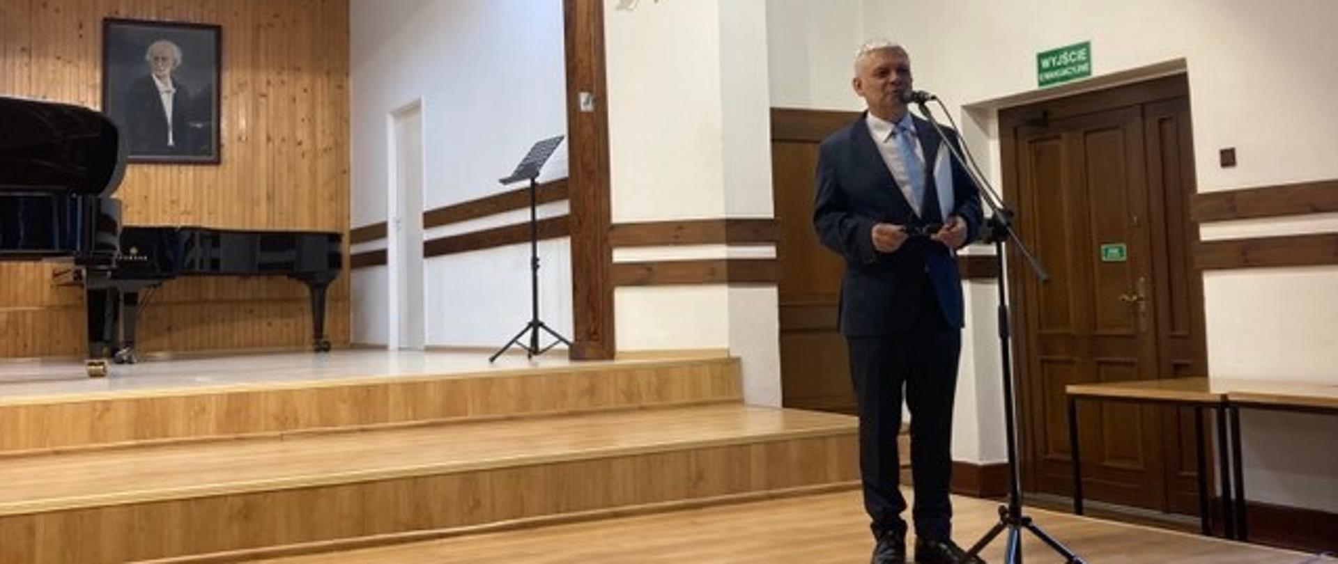 Pan Dyrektor ubrany w garnitur stoi i na scenie i mówi do mikrofonu.