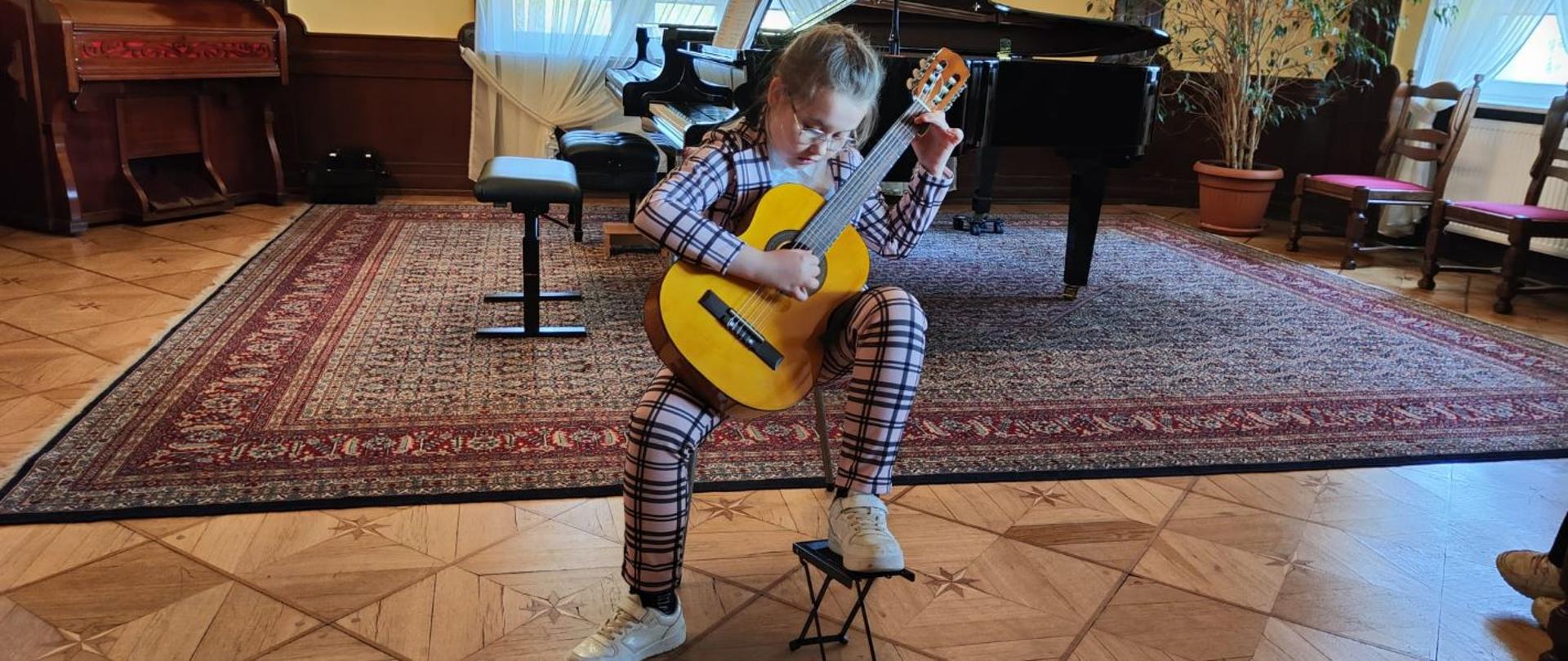 Zdjęcie kolorowe. Dziewczynka w okularach i kraciastym garniturze siedzi na krześle, grając na gitarze klasycznej. Jej lewa stopa spoczywa na małym podnóżku gitarowym. W tle duży, czarny fortepian skrzydłowy z otwartą klapą i nutami na pulpicie, stojący na wzorzystym dywanie. Po lewej stronie drewniane pianino lub fisharmonia, a po prawej roślina doniczkowa oraz krzesła. Pomieszczenie ma drewniany parkiet i jasne ściany z oknami zasłoniętymi firankami.