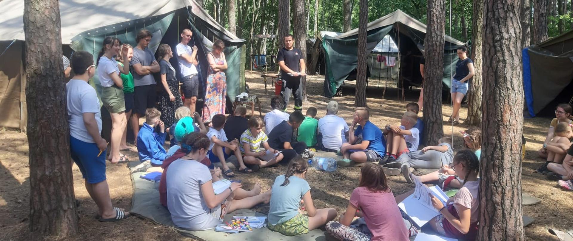 Młodzież stoi i siedzi na materacach w lesie. W tle widoczne namioty.