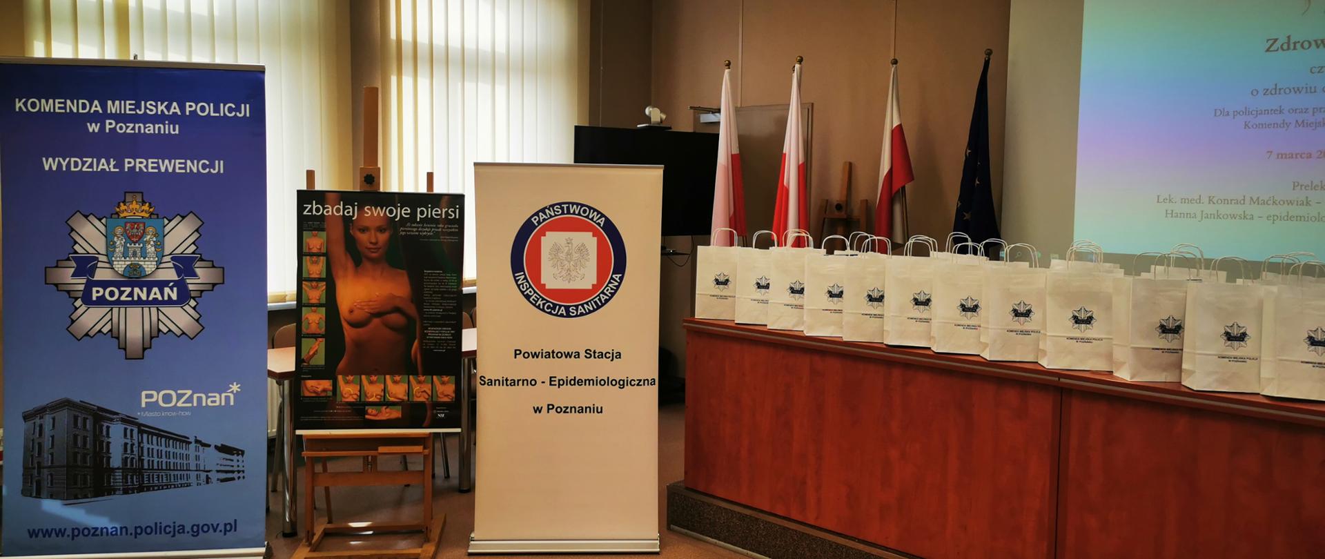 Na sali wykładowej stoją banery Powiatowej Stacji Sanitarno-Epidemiologicznej w Poznaniu oraz Komendy Miejskiej Policji w Poznaniu. Na stelażu plakat dot. samobadania piersi.