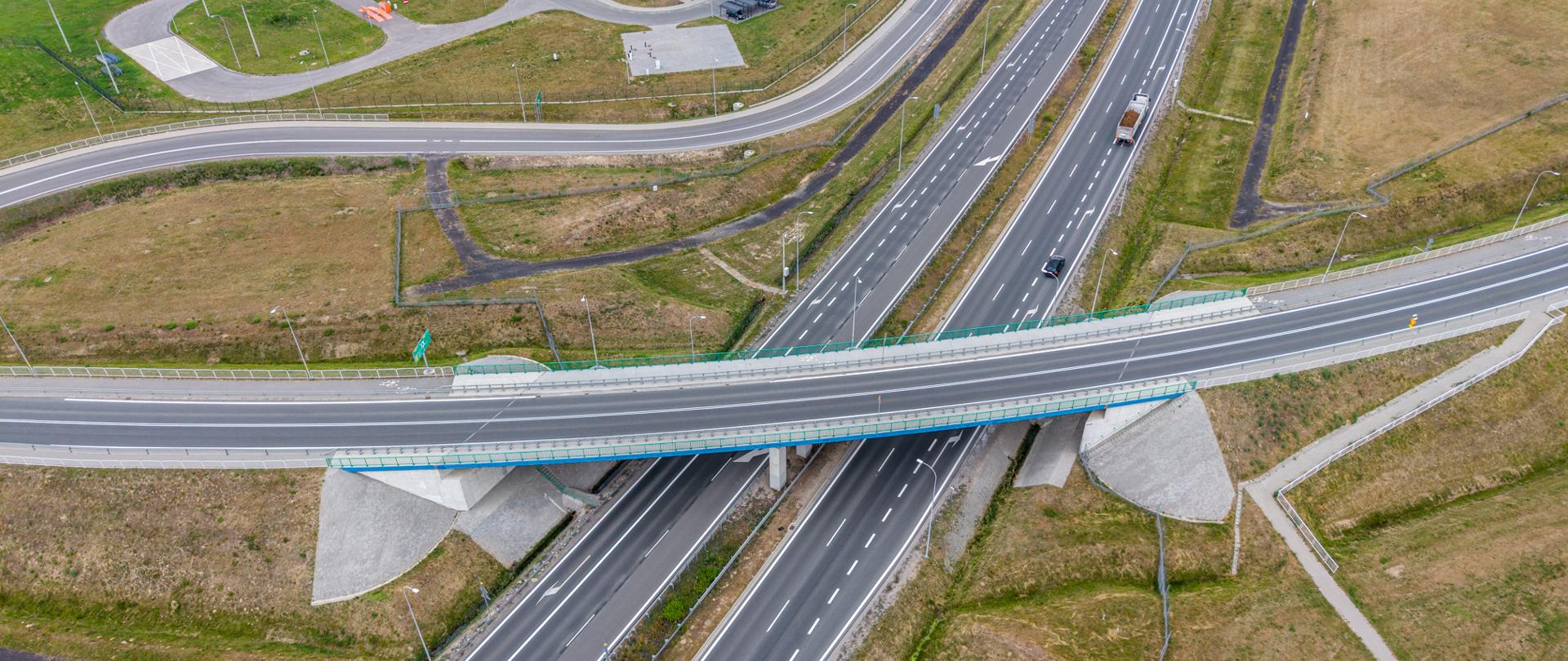 Zdjęcie lotnicze przedstawia nowoczesny węzeł drogowy z dwujezdniową trasą szybkiego ruchu. Nad główną drogą przebiega wiadukt, a wokół widoczne są łącznice umożliwiające wjazd i zjazd. W pobliżu znajduje się budynek zaplecza drogowego z parkingiem i placem sprzętowym. Otoczenie stanowią głównie trawiaste tereny oraz pola uprawne. Na jezdni widać pojedyncze pojazdy.