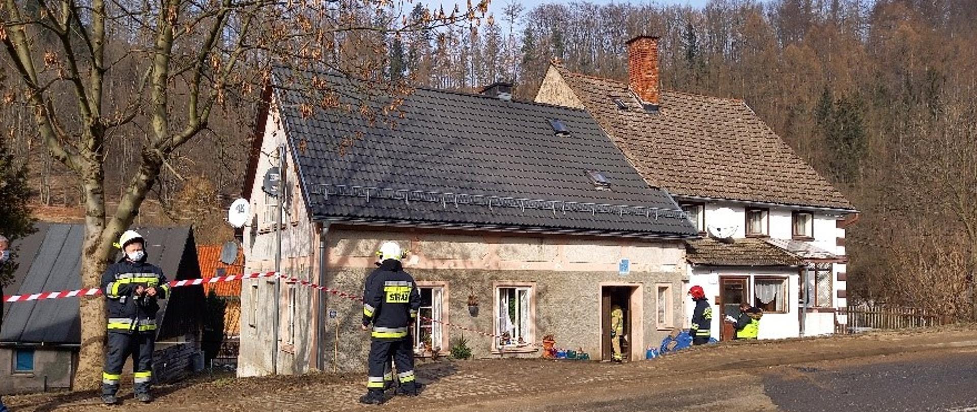 Murowany budynek, otaśmowany. Strażacy w ubraniu specjalnym. 