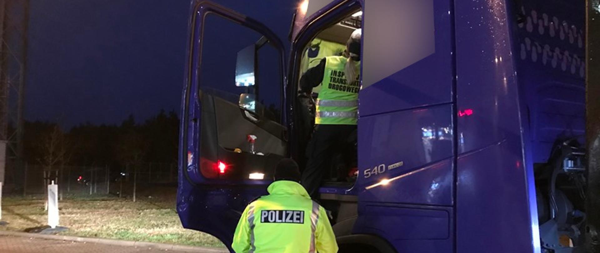 Inspektor w kabinie ciężarówki stoi na schodkach i pobiera dane z tachografu. Obok ciągnika siodłowego stoi niemiecki policjant w odblaskowej kurtce.