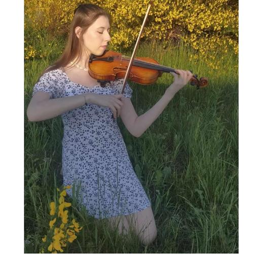 Zdjęcie kolorowe. Dziewczyna w białej sukience w czarne kropki. Gra na skrzypcach na ukwieconej łące.