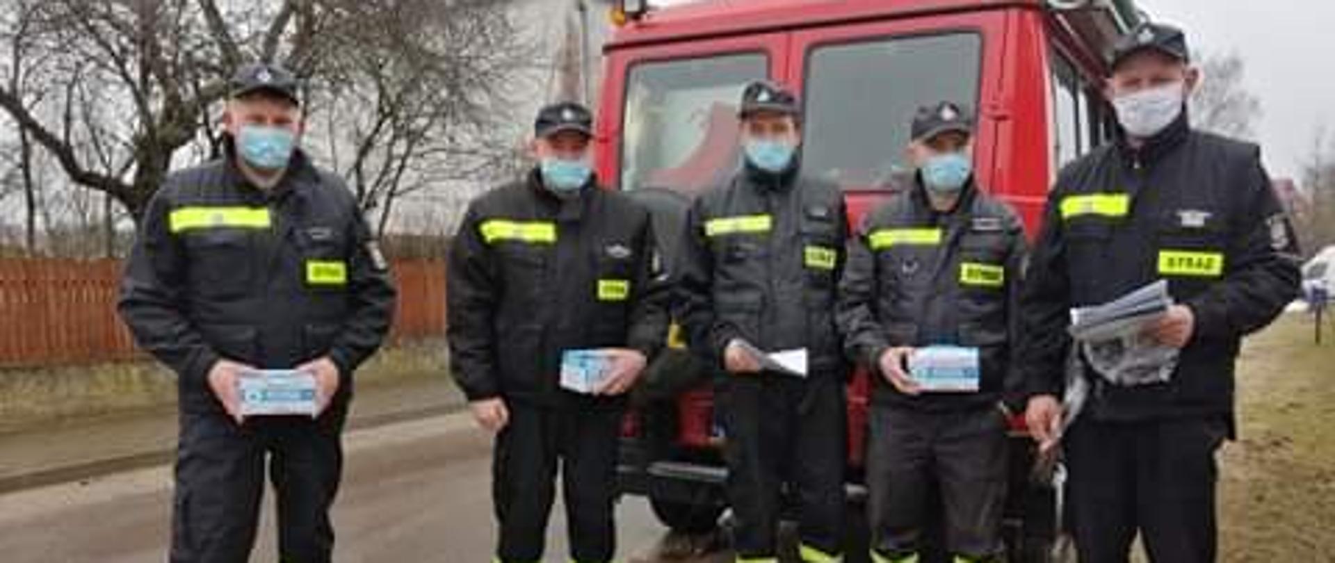 Zdjęcie przedstawia strażaków OSP na tle samochodu pożarniczego którzy trzymają maseczki jednorazowe oraz ulotki dotyczące szczepień