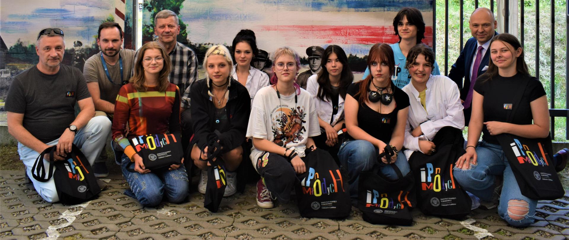 Zdjęcie pozowane. Wszyscy znajdują się w pozycji klęczącej i przysiadu. Na fotografii jest: Krzysztof Dąbek (dyr. PLSP w Lublinie), Robert Derewenda (dyr. IPN w Lublinie), nauczyciele: Tomasz Banaszkiewicz, Ryszard Paluch. Część osób trzyma w rękach czarne, materiałowe torby z kolorowymi napisami IPN Młodych.