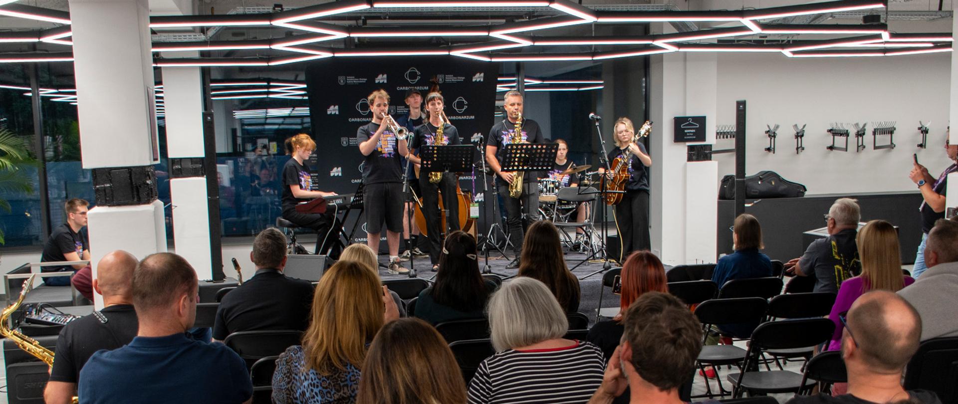 Siedmiu muzyków gra na scenie. Z przodu widoczna od tyłu publiczność.