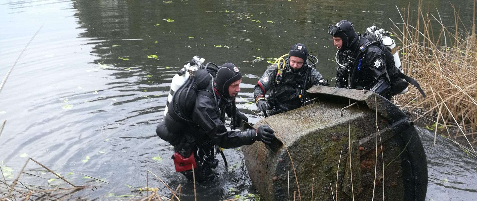 Zdjęcie przedstawia trzech strażaków - nurków którzy wydobywają na brzeg wielki metalowy pojemnik.