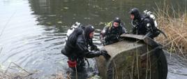 Zdjęcie przedstawia trzech strażaków - nurków którzy wydobywają na brzeg wielki metalowy pojemnik.