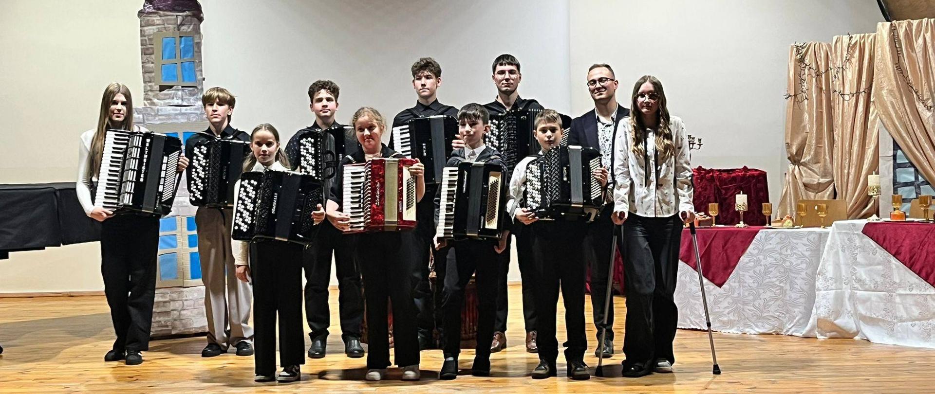 Ujęcie z bliska na grupę młodych artystów – dziewcząt i chłopców – pozujących z akordeonami na scenie. Po lewej stronie stoi makieta szarej wieży, po prawej stół z białym i czerwonym materiałem oraz dekoracyjne zasłony. Uczestnicy są uśmiechnięci i patrzą w stronę obiektywu, prezentując swoje instrumenty (w tym jeden akordeon z czerwonym miechem).