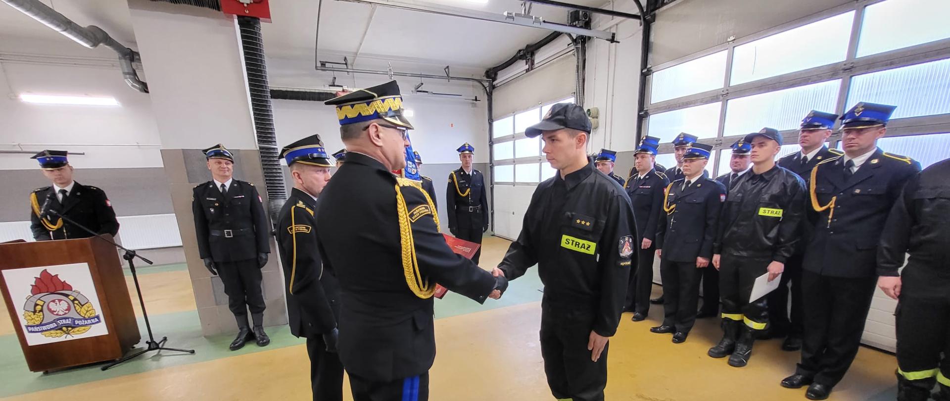 nadbrygadier Jacek Kaczmarek wraz ze starszym brygadierem Maciejem Suchomskim ubrany w mundur galowy gratuluje stojącemu naprzeciw aspirantowi Łukaszowi Kriegerowi ubranemu w mundur koszarowy, w tle poczet sztandarowy oraz strażacy ubrani w mundury galowe i koszarowe