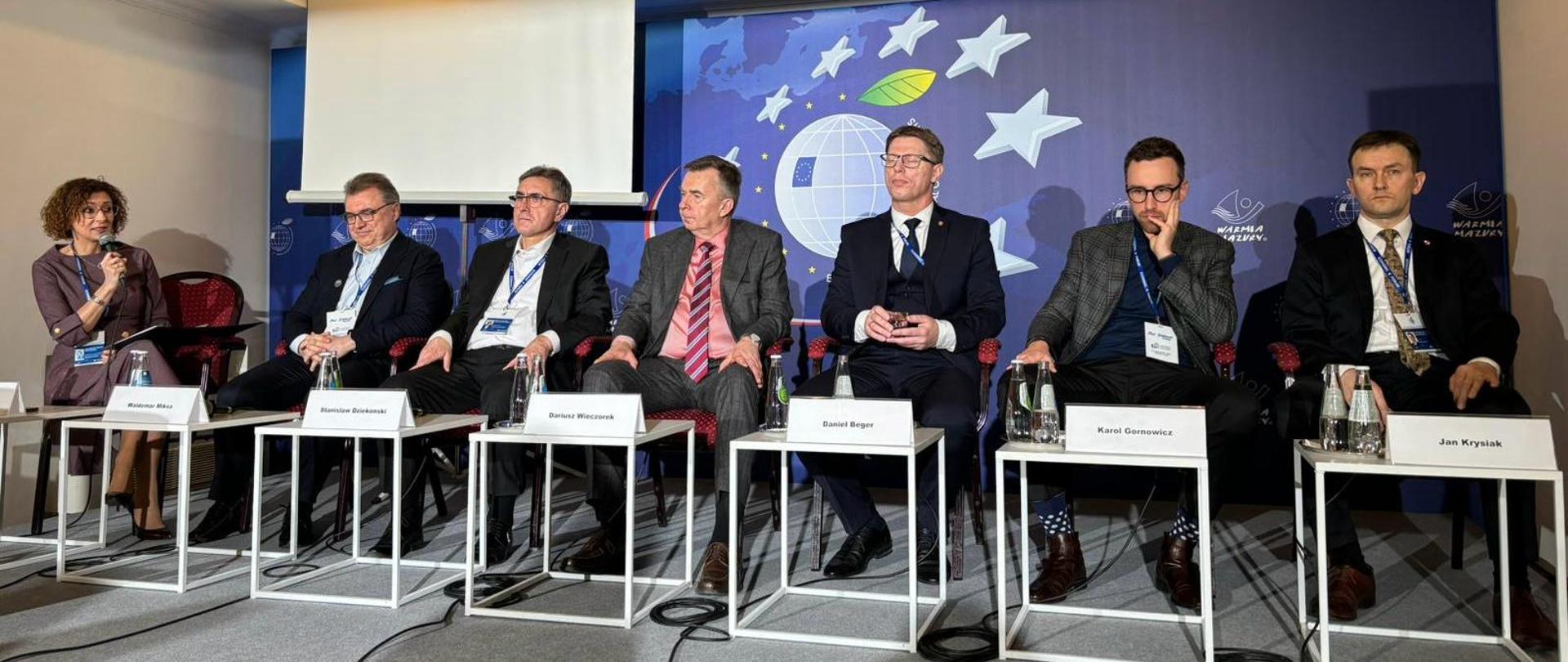 Pod ścianą przy małych białych stolikach siedzi sześciu mężczyzn, pośrodku minister Wieczorek, z boku na fotelu siedzi kobieta i mówi do mikrofonu.