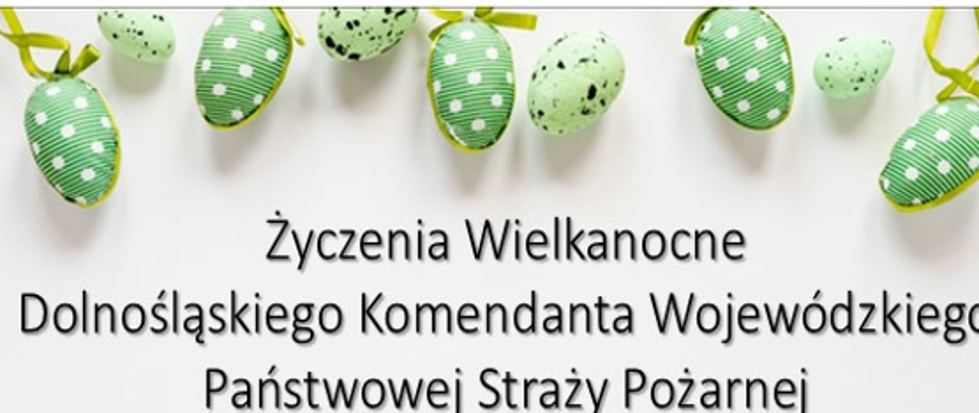 Grafika z życzeniami wielkanocnymi na białym tle. W centralnej części znajduje się czarny napis: „Życzenia Wielkanocne Dolnośląskiego Komendanta Wojewódzkiego Państwowej Straży Pożarnej”. Górną krawędź obrazu zdobi rząd zielonych pisanek – część gładka w czarne plamki, a część owinięta wstążkami w białe paski i kropki.