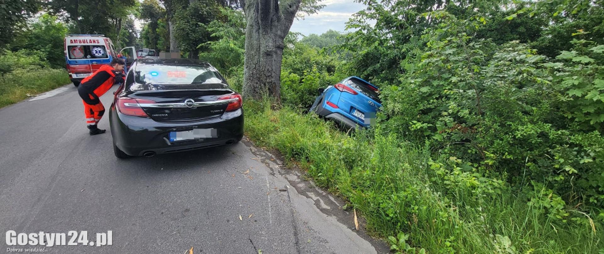 Samochód leży w rowie. Na drodze stoi samochód osobowy. W tle stoi karetka pogotowia.