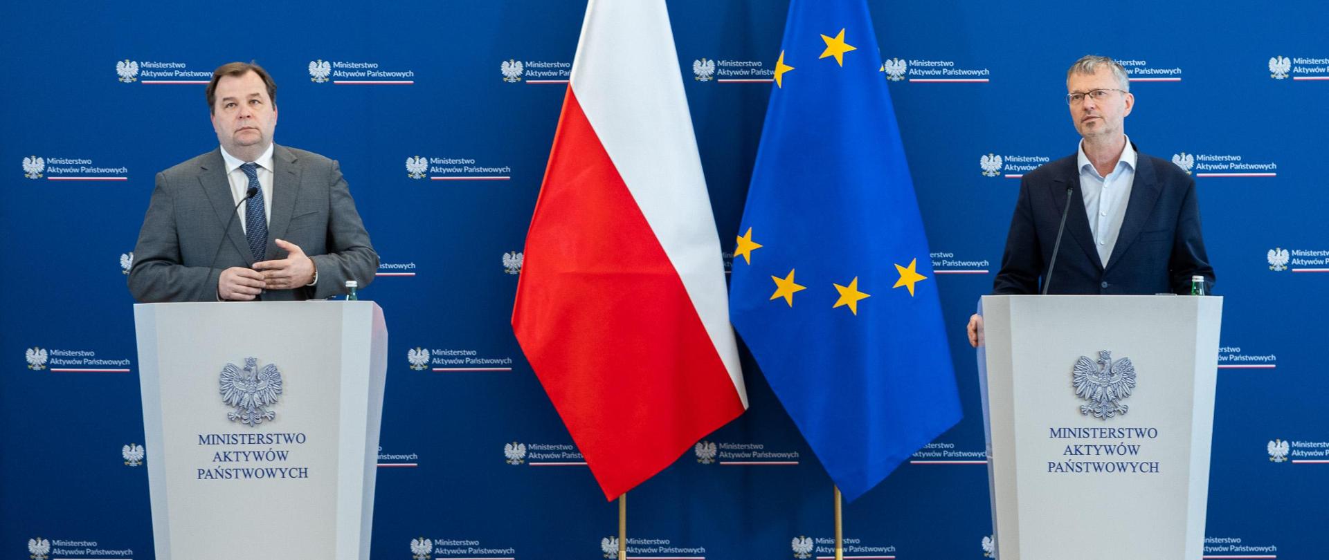 Na zdjęciu dwie osoby w garniturach stojące przy mównicach. Po środku flagi: Polski i Unii Europejskiej. W tle ścianka z logotypami Ministerstwa Aktywów Państwowych. 
