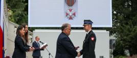 Centralnie na pierwszym planie Minister Andrzej Dera wręcza strażakowi w mundurze wyjściowych odznaczanie. Po lewej stronie dwie kobiety, w tle mężczyzna przy mikrofonie. W tle telebim z wizerunkiem odznaczania i napisem: Krzyż zasługi za dzielność.