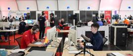 Widok z boku na wnętrze hali, przy stolikach przed komputerami siedzą ludzie w różnokolorowych koszulkach.
