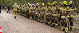Na zdjęciu widzimy grupę strażaków stojących w szeregu na leśnej drodze gruntowej. Są ubrani w pełne umundurowanie ochronne: kaski, rękawice oraz kombinezony z odblaskowymi pasami. Jeden ze strażaków, z napisem „STRAŻ” na plecach, oddala się od grupy w lewą stronę kadru, co może sugerować, że pełni rolę dowódcy lub wykonuje konkretne zadanie.
Otoczenie to gęsto zalesiony teren, a pogoda wydaje się pochmurna — typowe warunki dla ćwiczeń terenowych lub akcji ratowniczej. W tle widać kilka innych osób, które mogą być obserwatorami, koordynatorami lub członkami innych służb.
Cała scena wygląda na dobrze zorganizowaną — może to być część szkolenia, manewrów lub przygotowań do działań w terenie. Obecność wielu strażaków i ich ustawienie wskazują na gotowość i dyscyplinę.
