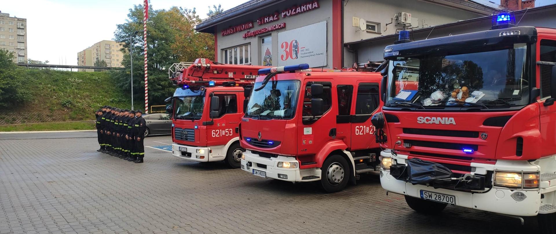 Zdjęcie przedstawia strażaków oddających hołd. Na pierwszym planie znajdują się samochody pożarnicze.