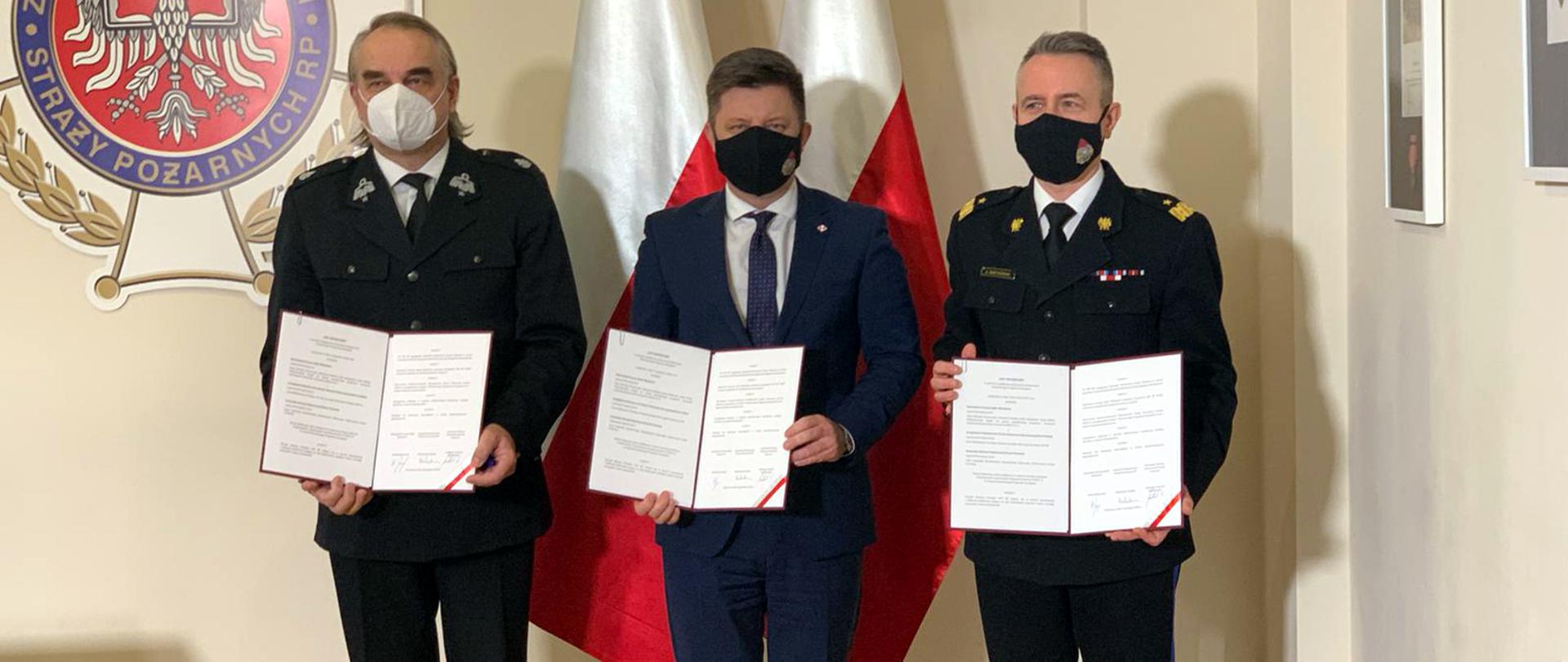 Zdjęcia przedstawia trzy osoby, które prezentują podpisany list intencyjny pomiędzy Kancelarią Prezesa Rady Ministrów, Zarządem Głównym ZOSP RP oraz Komendą Główną PSP. W tle stoja dwie Flagi Rzeczypospolitej Polskiej. Po lewej stronie zdjęcia u góry herb ZOSP RP.
